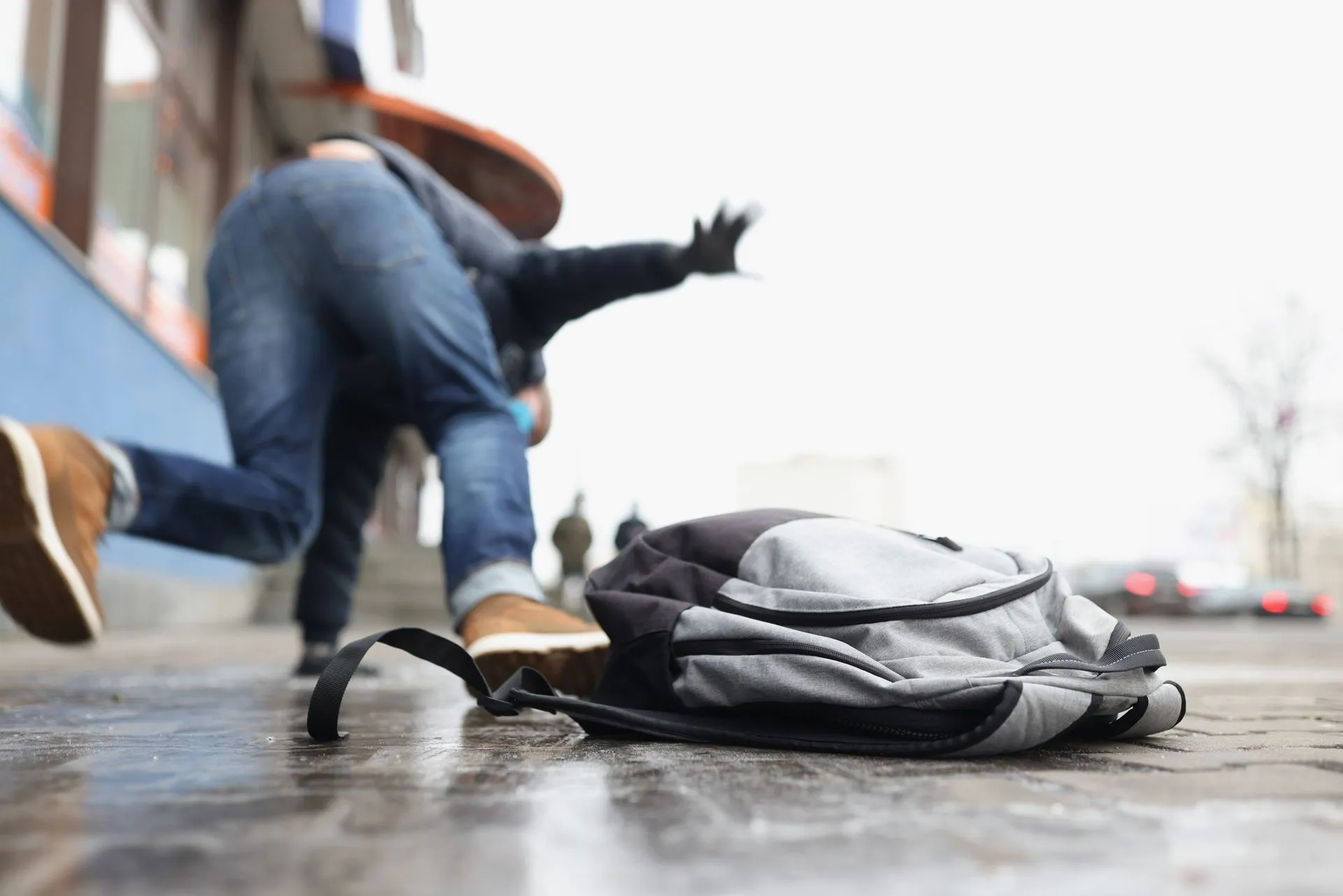 Person tripping on a wet sidewalk, backpack lying on the ground, city background.