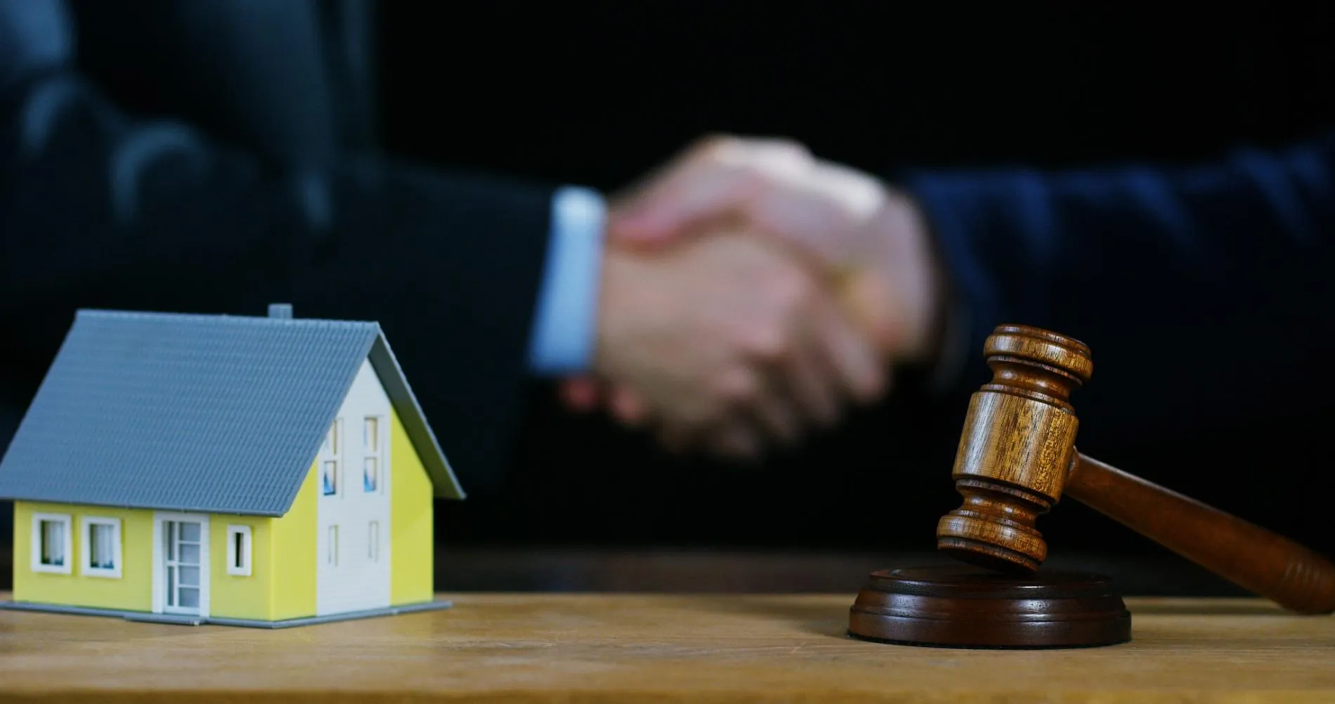 A miniature house model and wooden gavel on a table with two people in suits shaking hands in the blurred background.