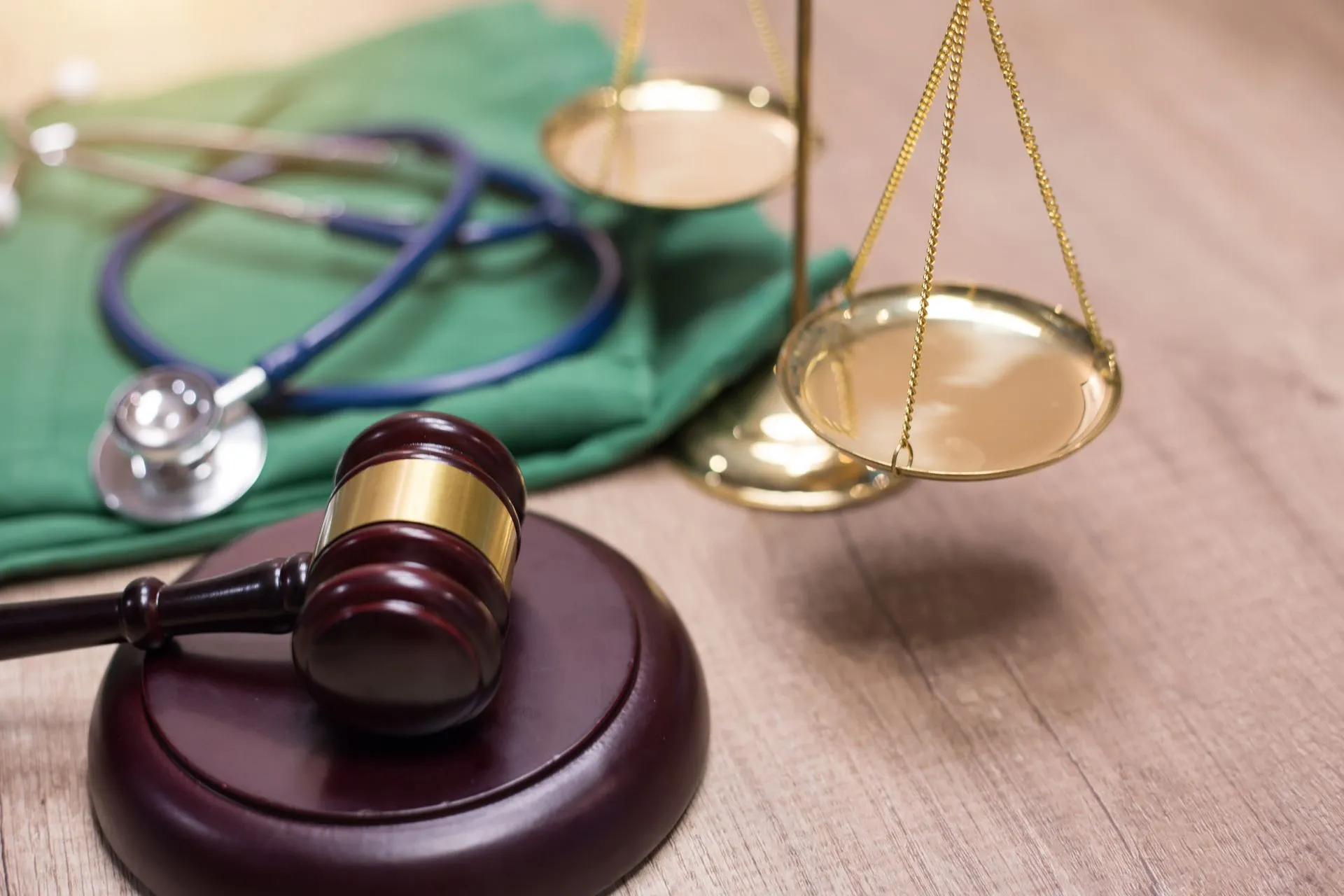 Wooden gavel and brass scales of justice resting on a table next to a stethoscope and green medical scrubs.